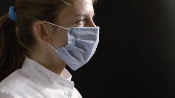 The female doctor or nurse slowly turns her head and looks into the camera against the black background. How Doctors Work. - Imágenes, Vídeo