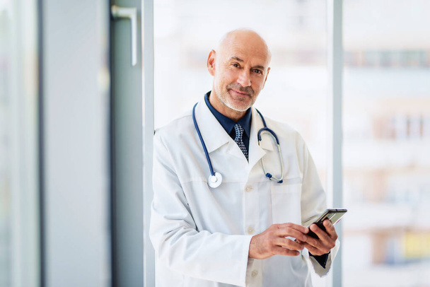 Portrait of a male doctor using mobile phone and texting while standing in the corridor of the hospital. - Photo, image