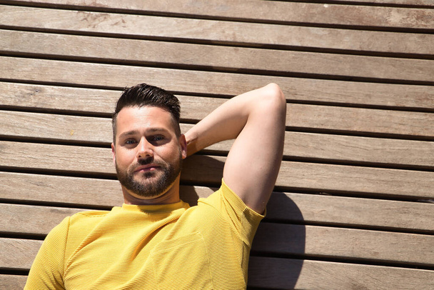 Portrait of handsome young man, with blue eyes and yellow t-shirt, lying on a wooden slatted surface smiling happily. Concept beauty, fashion, happiness, trend. - Φωτογραφία, εικόνα