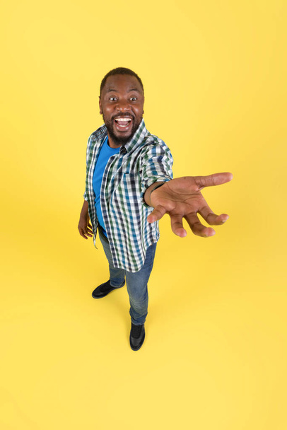 Funny Black Guy Showing Invisible Object Shouting Stretching Hand To Camera Standing In Studio Over Yellow Background. Man Holding And Advertising Product. Vertical - Photo, Image