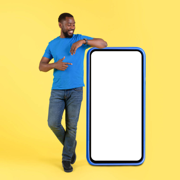 Positive Black Male Pointing At Big Phone With Blank Screen Advertising Mobile Application Over Yellow Studio Background. Check This App Concept. Square, Full Length - Photo, Image