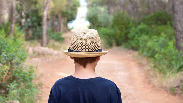 Boy with straw hat and dark blue t-shirt walking towards the beach between a forest of pine trees - Valokuva, kuva