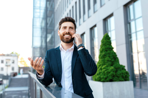 Cheerful young european businessman with beard in suit calling by phone on balcony of modern office building, outdoor. Ceo manager uses gadget for business remotely, talking with client and meeting - 写真・画像