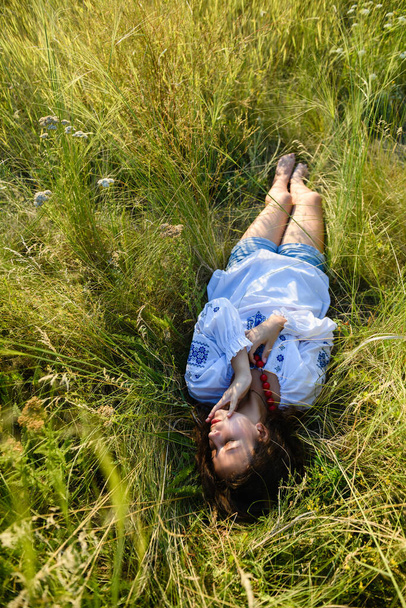 A teenage girl lies in a tall meadow grass. The girl is dressed in a national Ukrainian shirt - vyshyvanka. Top view. - Zdjęcie, obraz