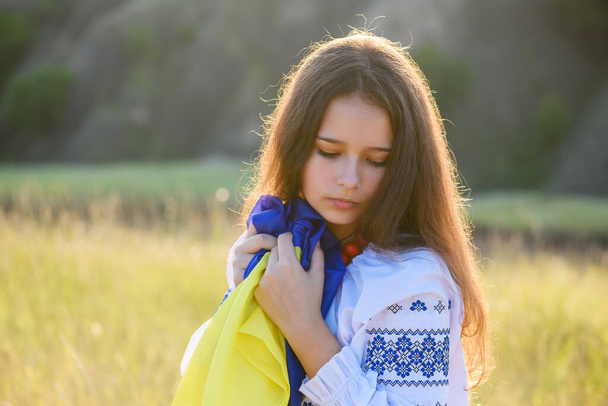 A sad teenage girl holds a Ukrainian flag to her chest. The girl is dressed in Ukrainian national clothes - vyshyvanka. Soft backlight at sunset. - Fotó, kép