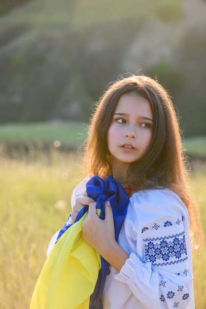 A sad teenage girl holds a Ukrainian flag to her chest. The girl is dressed in Ukrainian national clothes - vyshyvanka. Soft backlight at sunset. - Foto, Bild