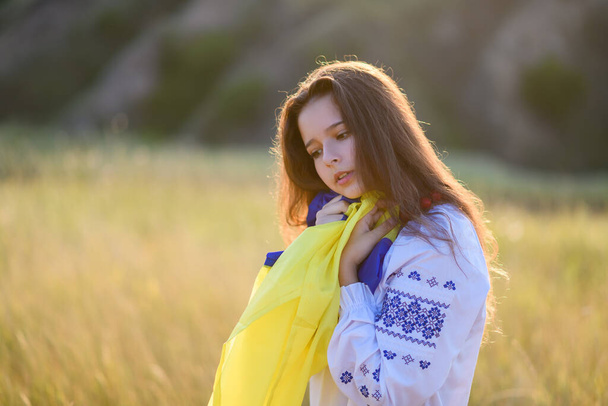 A sad teenage girl holds a Ukrainian flag to her chest. The girl is dressed in Ukrainian national clothes - vyshyvanka. Soft backlight at sunset. - Foto, afbeelding