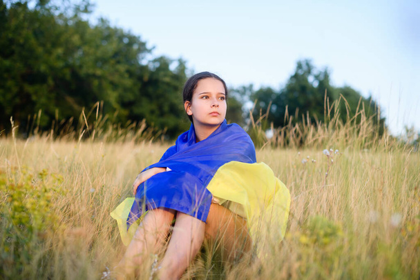 A teenage girl sits in tall wild grass on the slope of a ravine with a Ukrainian flag on her shoulders. - Fotó, kép