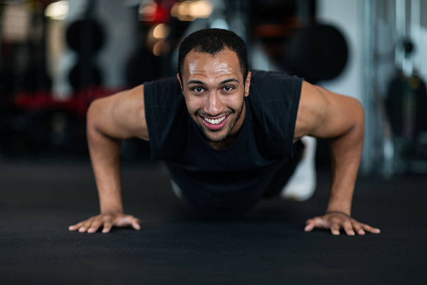 Smiling Handsome Black Man Making Push Up Exercises While Training At Gym, Young Muscular African American Guy Exercising In Modern Sport Club, Working Out On Floor, Closeup Shot With Copy Space - Photo, Image