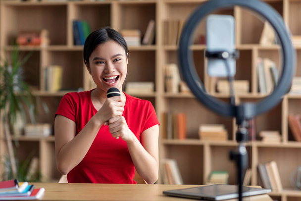 Cheerful pretty young japanese female superstar singing at microphone, shooting blog on smartphone in room interior. Hobbies and modern technology, karaoke and entertainment during covid-19 outbreak - Photo, Image