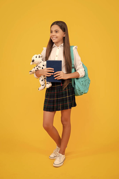 счастливый ребенок с рюкзаком игрушки и книгой в школьной форме в полный рост на желтом фоне, учеба. - Фото, изображение