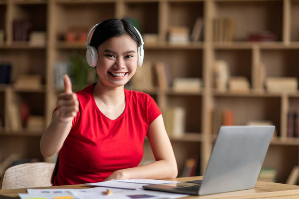 Glad young asian woman with down syndrome in headphones with laptop point finger at camera in office interior. Your turn, education at home, study and social distancing, ad and offer during covid-19 - Φωτογραφία, εικόνα