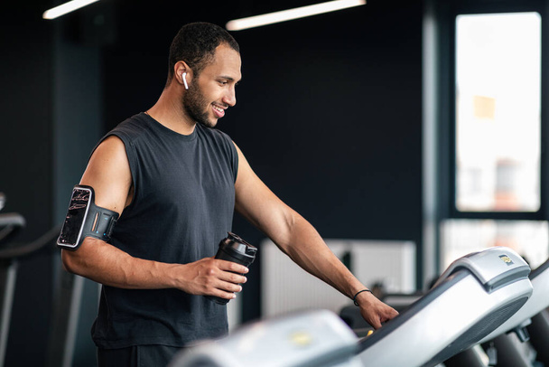Portrait Of Happy Black Male Athlete Exercising On Treadmill At Gym, Smiling Young African American Man Holding Fitness Shaker Bottle And Selecting Running Mode On Control Panel, Copy Space - Photo, image