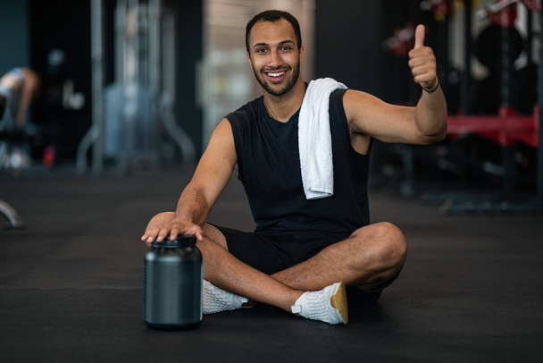 Fitness Nutrition. Smiling African American Male Athlete Demonstrating Container With Whey Protein Powder And Showing Thumb Up, Sporty Black Guy Recommending Workout Supplement While Posing In Gym - Fotó, kép