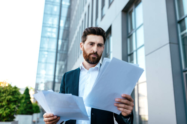 Busy confident young businessman with beard in suit works with documents near office building, outdoor. Leadership and project analysis, ceo manager, boss and contract, develop marketing strategy - Foto, immagini