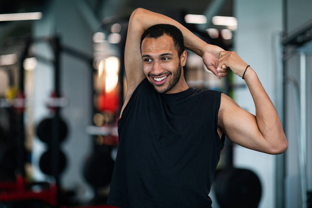 Portrait Of Athletic Black Guy Stretching Arm Muscles At Gym, Young Sporty African American Man Warming Up Before Training In Modern Fitness Club, Enjoying Exercising, Closeup Shot With Copy Space - Foto, afbeelding