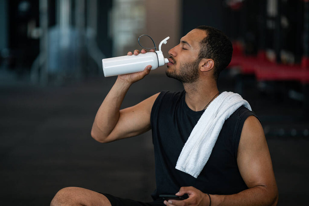 Workout Break. Sporty Young Black Man Drinking Water After Exercising At Gym, African American Male Athlete Resting In Modern Sport Club, Enjoying Refreshing Grink And Holding Smartphone, Closeup - Фото, изображение