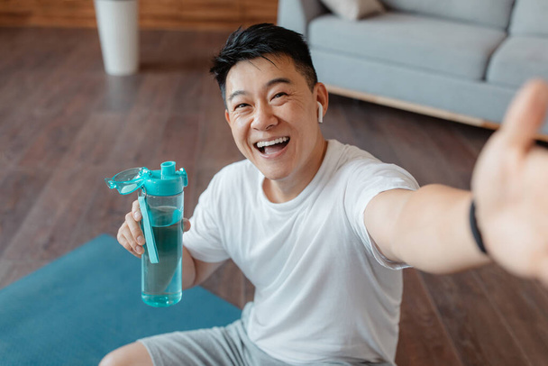 Happy mature asian man with bottle of water taking selfie on break from home workout, sitting on fitness mat and making photo of himself during domestic training - 写真・画像