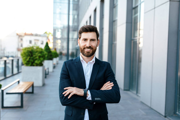 Leadership, ceo manager and boss. Smiling confident young attractive european man with beard in suit with crossed arms on chest near modern office building, outdoor. Work and business, ad and offer - Foto, Bild