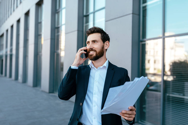 Happy busy confident handsome businessman with beard in suit works with documents and calls by smartphone near office building, outdoor. Ceo manager and conversation with client, boss and contract - Foto, Bild