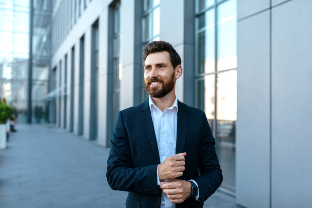 Smiling confident millennial attractive caucasian businessman with beard in suit looks at free space stands near modern office building, outdoor. Successful ceo manager and boss, business in city - Fotoğraf, Görsel