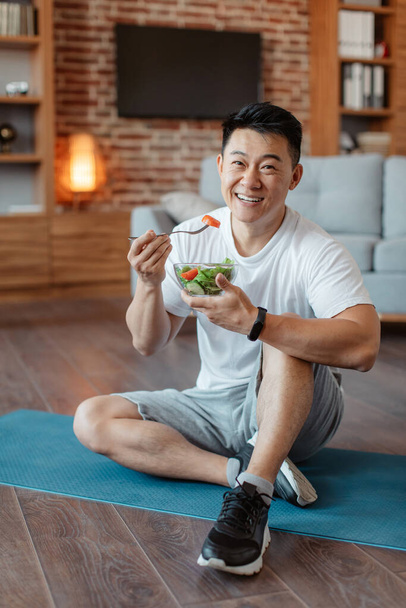 Healthy lifestyle concept. Happy middle aged asian man eating fresh vegetable salad, sitting on yoga mat after home workout and smiling at camera. Mature male keeping weight loss diet - Fotó, kép