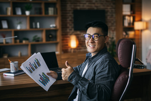Happy adult asian businessman in glasses working with charts and showing thumb up, thinking about online project in home office interior. Analyze statistic and approve successful business remotely - Foto, Imagen