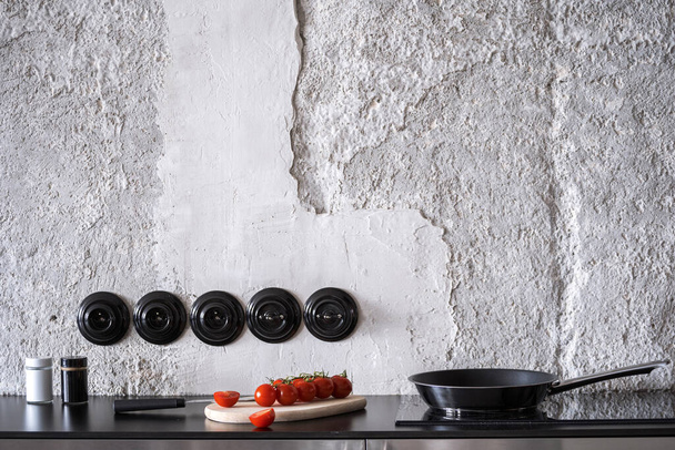 empty frying pan with handle on glass ceramic hob, red fresh tomatoes with knife on wooden board on black worktop at modern kitchen in apartment - 写真・画像
