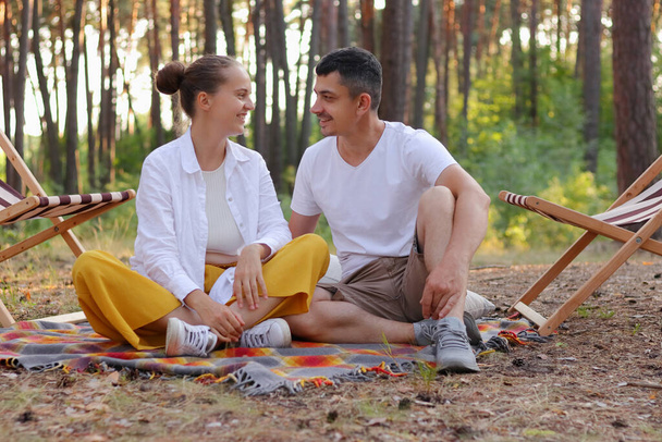 Outdoor shot of beautiful young couple sitting on a plaid in an summer park, looking at each other with love and gentle, expressing positive emotions, having vacation in the forest. - Foto, Imagem