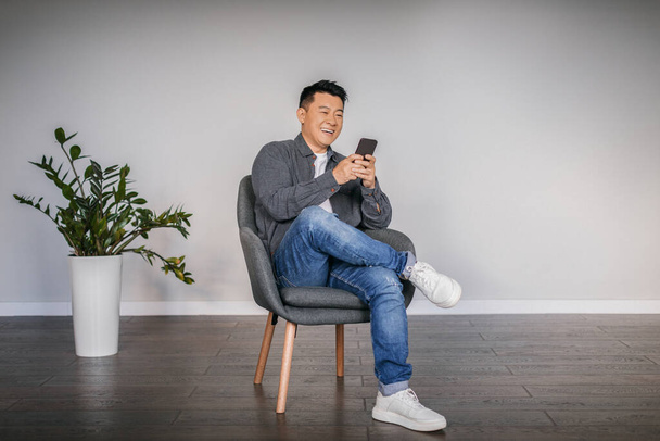 Happy adult asian man typing on smartphone sitting in armchair in minimalist living room interior, free space. Chatting in social networks and surfing in Internet, new application and great offer - Photo, Image