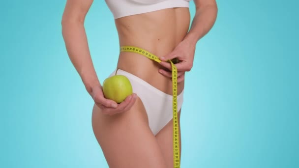 Healthy nutrition and slimming. Close up shot of unrecognizable fit lady measuring her figure with tape, holding organic green apple, blue studio background, slow motion - Materiał filmowy, wideo