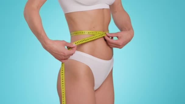 Weight Loss concept. Close up shot of unrecognizable slim lady measuring her waist with tape, calculating her slimming, blue studio background, slow motion - 映像、動画