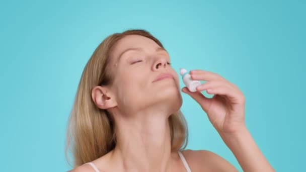 Runny nose treatment. Sick middle aged woman using nasal drops, getting treatment and smiling after relief effect, blue studio background, slow motion - Video