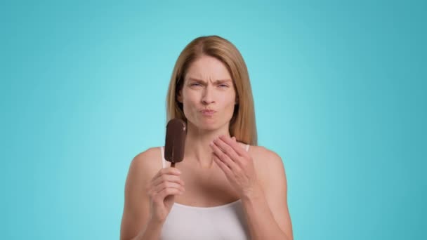Tooth sensitivity. Middle aged woman biting ice-cream and suffering from acute toothpain, touching her painful jaw, blue studio background, slow motion - Materiał filmowy, wideo