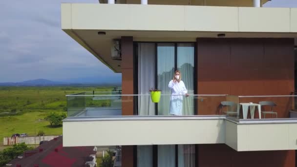 Yazın güneşli bir günde otelin balkonunda beyaz önlüklü ve bir fincan kahve içen mutlu sarışın bir kadının robot görüntüsü. - Video, Çekim