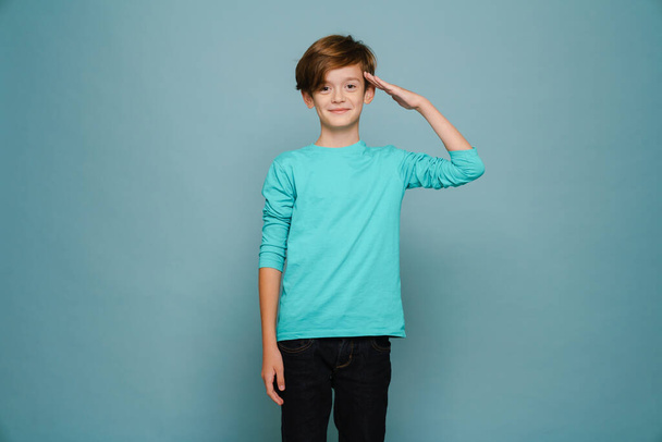 Ginger white boy wearing long sleeve smiling and saluting at camera isolated over blue wall - Fotografie, Obrázek