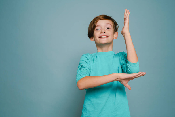 Ginger boy wearing long sleeve smiling and stretching hand upward isolated over blue wall - 写真・画像