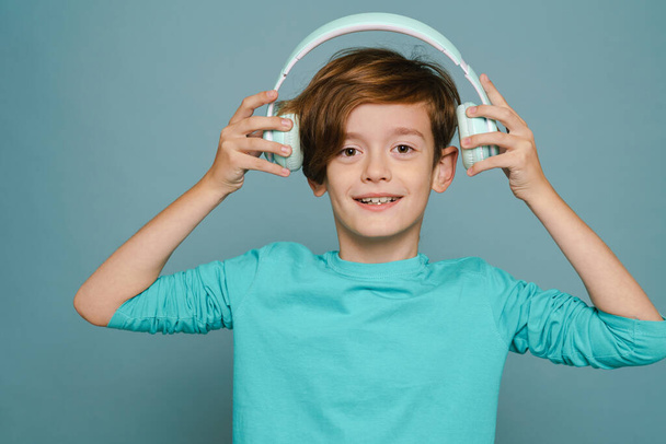 Ginger white boy smiling while listening music with headphones isolated over blue wall - Valokuva, kuva