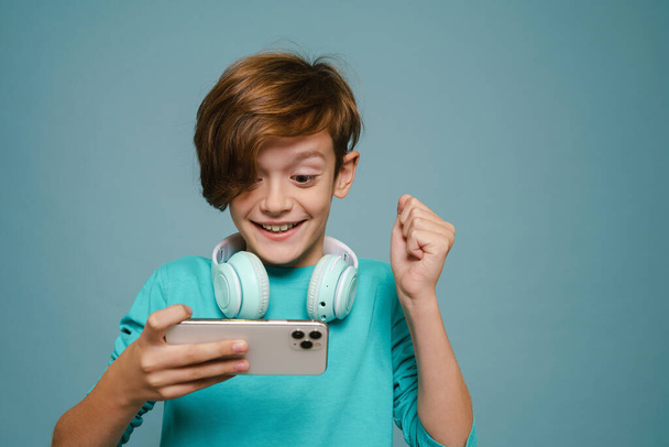 Ginger white boy gesturing while playing online game on cellphone isolated over blue wall - 写真・画像