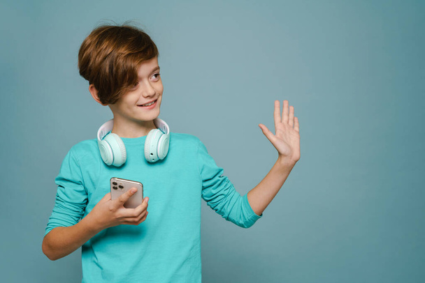 Ginger white boy with headphones gesturing and using cellphone isolated over blue wall - Valokuva, kuva