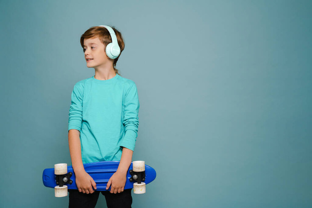 Ginger white boy in headphones posing with skateboard isolated over blue wall - Fotó, kép