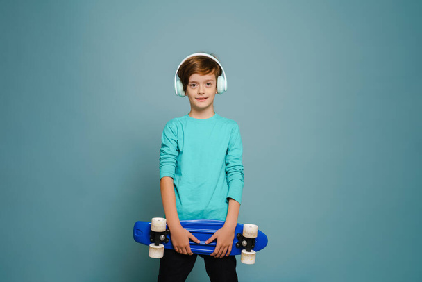 Ginger white boy in headphones smiling while posing with skateboard isolated over blue wall - Photo, image