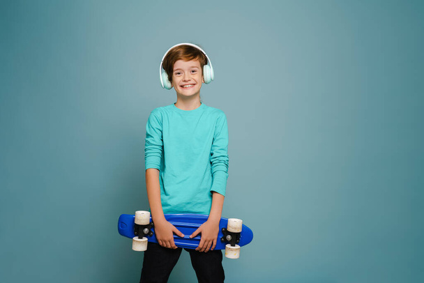 Ginger white boy in headphones smiling while posing with skateboard isolated over blue wall - Foto, imagen