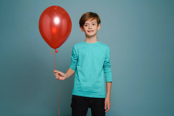 Ginger white boy smiling while posing with red balloon isolated over blue wall - Foto, afbeelding