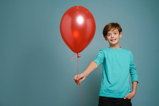 Ginger white boy smiling while posing with red balloon isolated over blue wall - Фото, зображення