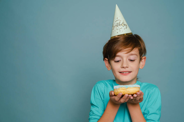 Ginger boy wearing party cone smiling while posing with doughnut isolated over blue wall - Foto, afbeelding