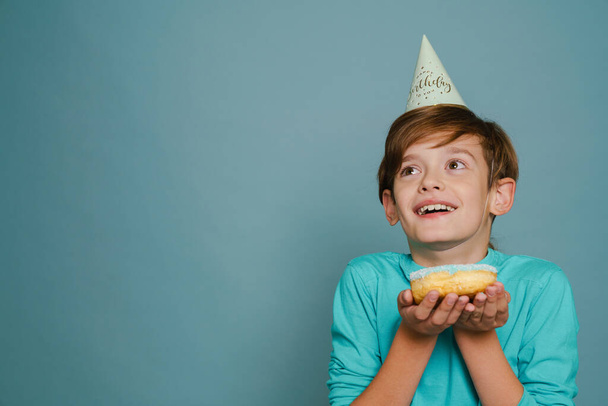 Ginger boy wearing party cone smiling while posing with doughnut isolated over blue wall - Zdjęcie, obraz