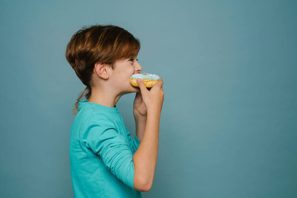 Ginger white boy wearing long sleeve eating doughnut isolated over blue wall - 写真・画像