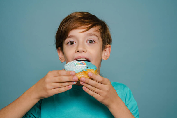 Ginger white boy wearing long sleeve eating doughnut isolated over blue wall - Фото, изображение
