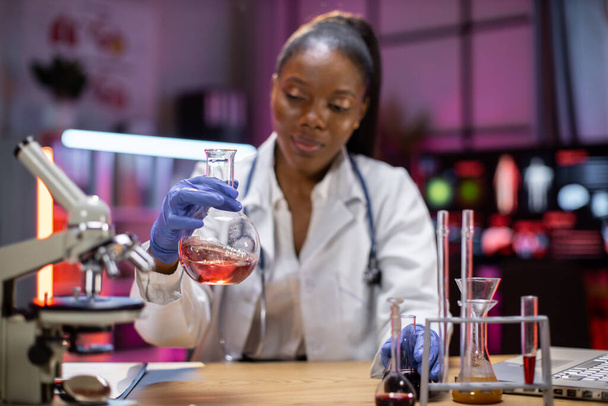 Positive african american female scientist or medical in lab coat holding test tube with reagent, laboratory glassware containing chemical liquid. Microscope, Biochemistry laboratory research. - Zdjęcie, obraz